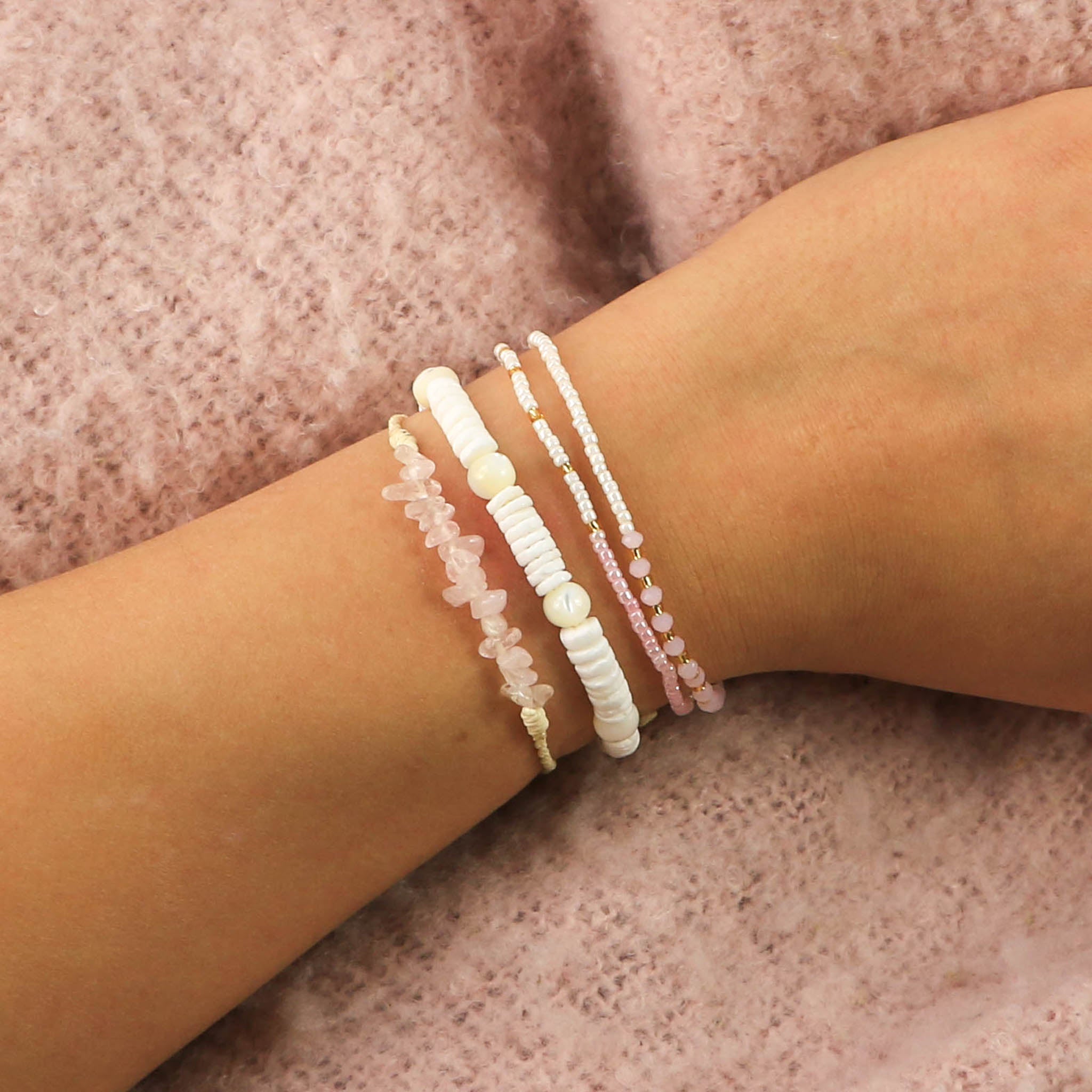 A model wearing our limited edition Galentines (Valentines) bracelet set on a pink background. A white, pink and gold double layer beaded bracelet, a white puka shell and shimmering troca shell bead bracelet, and our Love Is Here rose quartz gemstone bracelet.