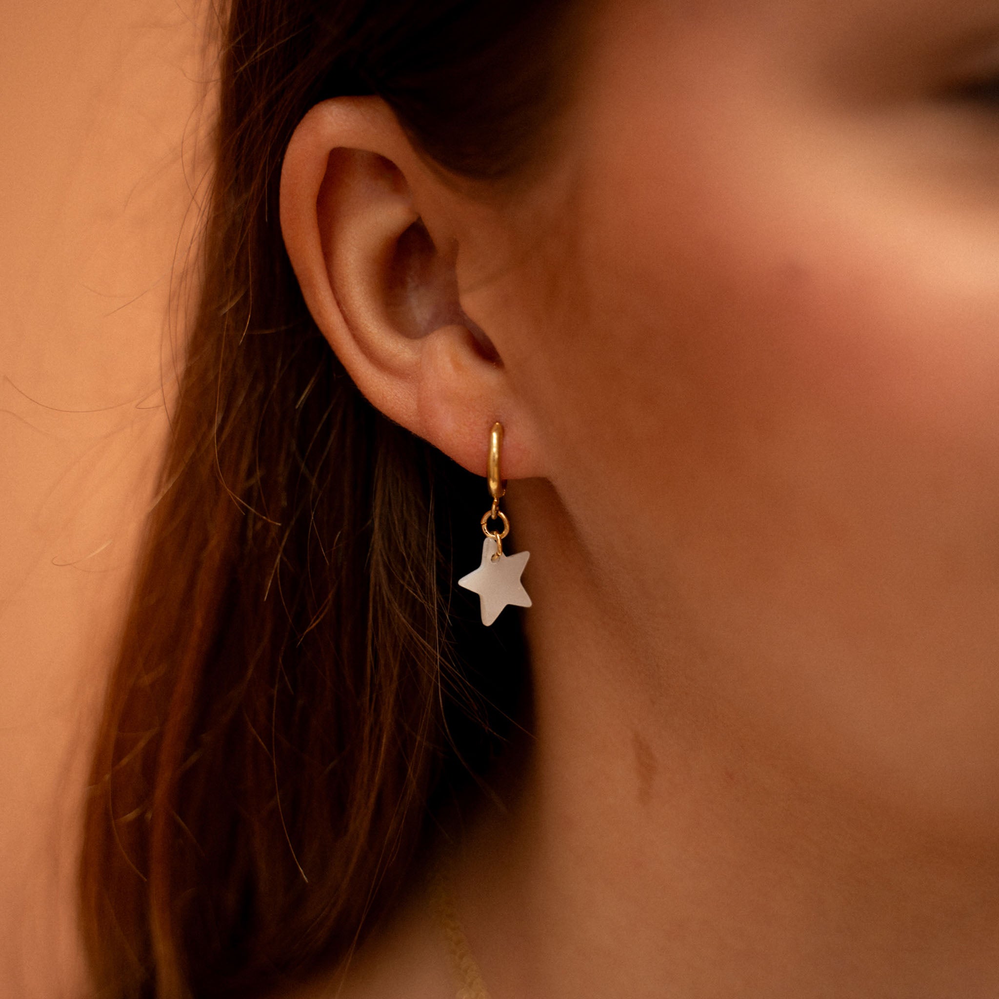 Celestial inspired star charm earrings, gold-plated copper alloy huggie hoops adorned with a star shaped, flat mother-of-pearl charm. Pictured on a models ear.