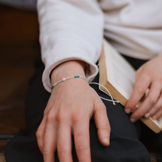 A persons wrist wearing a dainty beaded bracelet with blue toned, white and turquoise beads next to an open book on their lap