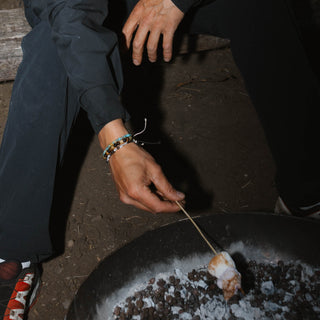 Person roasting marshmallows on an open fire in a dark setting, wearing beaded bracelets