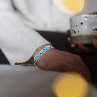 Close up of a person holding a mug in one hand, and a turquoise and white beaded bracelet on the other, with blurred fairy lights in the foreground