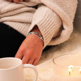Person wearing a beige sweater and two bracelets on the wrist, with a mug and candle in the foreground
