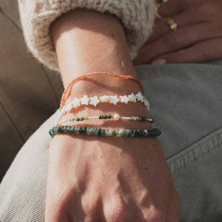 A close-up of a person's wrist wearing four layered beaded bracelets. The bracelets include a copper-orange beaded bracelet, a white star-shaped bead bracelet, a thin green and white beaded bracelet, and a dark green marbled stone bracelet.